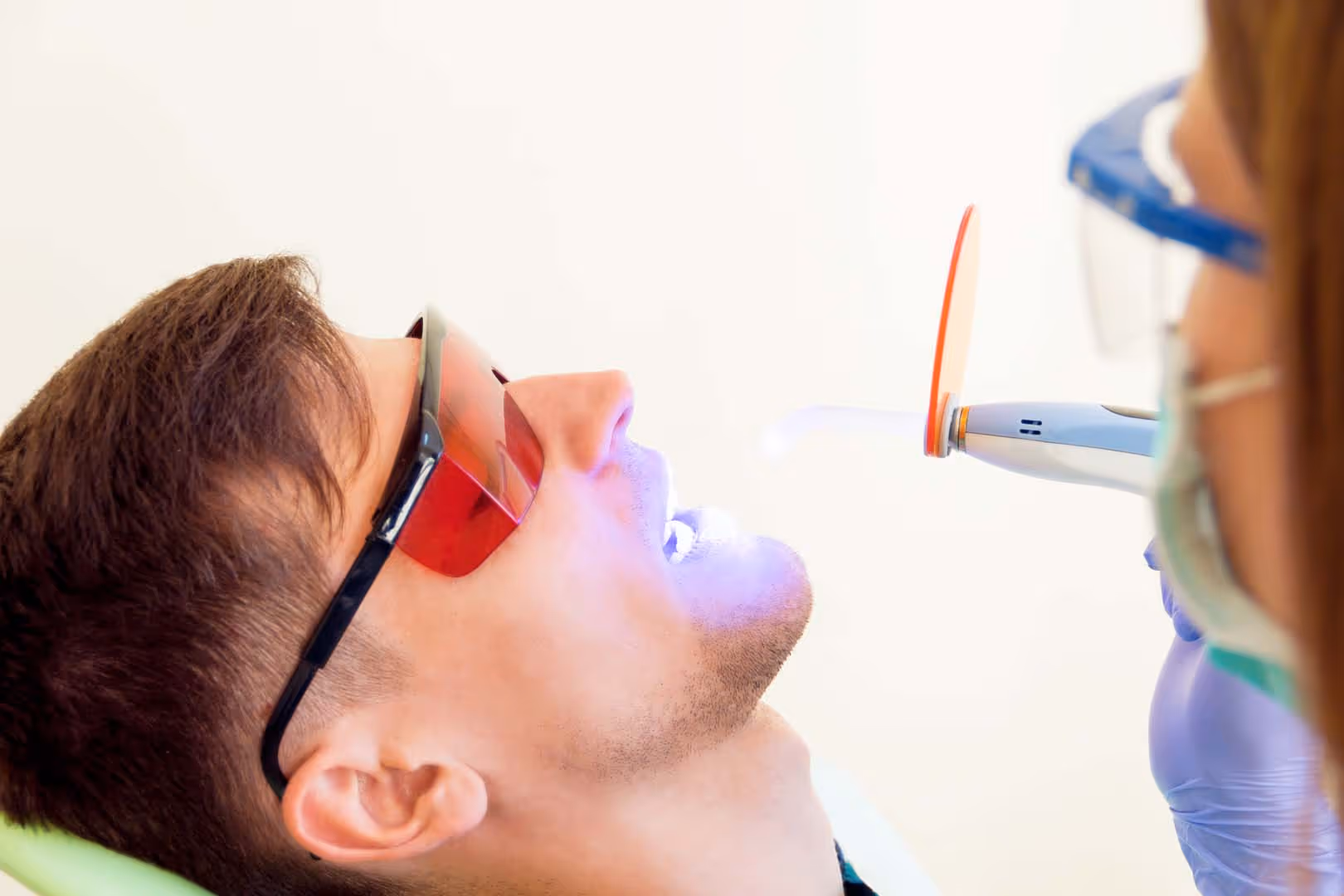 Young woman female dentist in medical glasses and mask treatments teeth of man male patient in red uv glasses by uv equipment tool implanting medical sealants